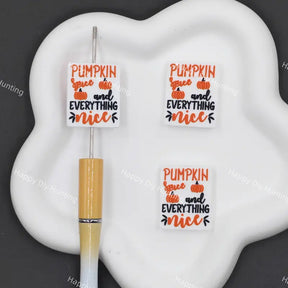 Three white square decorative stickers with the text 'PUMPKIN spice and EVERYTHING nice' in orange and black, displayed on a white cloud-shaped tray next to a gold and white pen on a dark gray background.