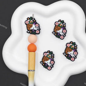 Four embroidered patches featuring a white cat in a basket decorated with flowers and a green hat, displayed on a white cloud-shaped tray next to a wooden bead pen.