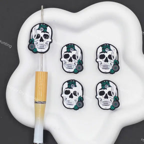 Four white skull-shaped charms with black outlines, green floral accents, and gray beads are arranged on a white cloud-shaped tray next to a yellow-and-white pen on a dark gray background.