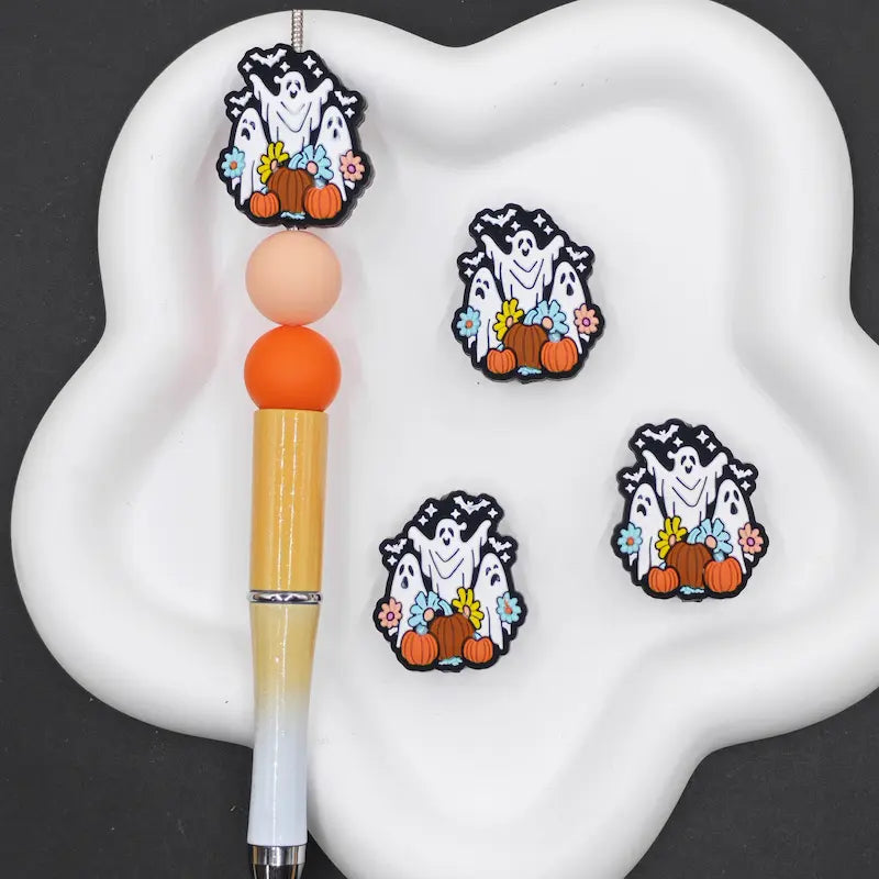 Three Halloween-themed ghost and pumpkin charms are displayed on a white cloud-shaped paint palette next to a dual-tone paint stick. The charms feature white ghosts with black outlines, orange pumpkins, and colorful flowers.
