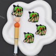 Four cartoon skull-and-crossbones charms displayed on a white cloud-shaped tray, alongside a decorative pen on a dark gray background.