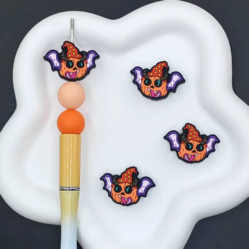 Three small cartoon bat charms with orange hats and purple wings are displayed on a white cloud-shaped tray, alongside a pen used for scale.