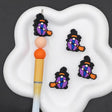 Three small witch-themed charms with black hats, purple bodies, blue faces, and orange brooms are arranged on a white cloud-shaped tray next to a paintbrush on a dark background.