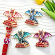 Four mixed color book winged dragon metal fancy beads with rhinestones displayed on a white wavy tray. Colors include red, blue, teal, and purple, each featuring a detailed dragon with enamel wings sitting on red books, plus a red rhinestone beadable pen. Perfect for DIY beadable pens and crafts.