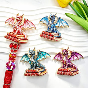 Four mixed color book winged dragon metal fancy beads with rhinestones displayed on a white wavy tray. Colors include red, blue, teal, and purple, each featuring a detailed dragon with enamel wings sitting on red books, plus a red rhinestone beadable pen. Perfect for DIY beadable pens and crafts.
