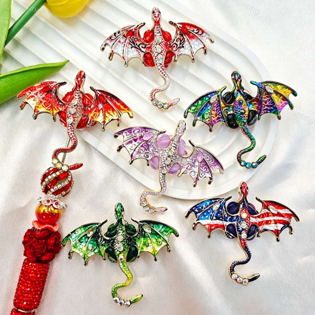 Five mixed color flying winged dragon metal fancy beads with rhinestones displayed on a white wavy tray. Colors include red, green, blue, purple, and rainbow, each featuring a detailed dragon with enamel wings and a rhinestone-studded body. Perfect for DIY beadable pens and crafts.