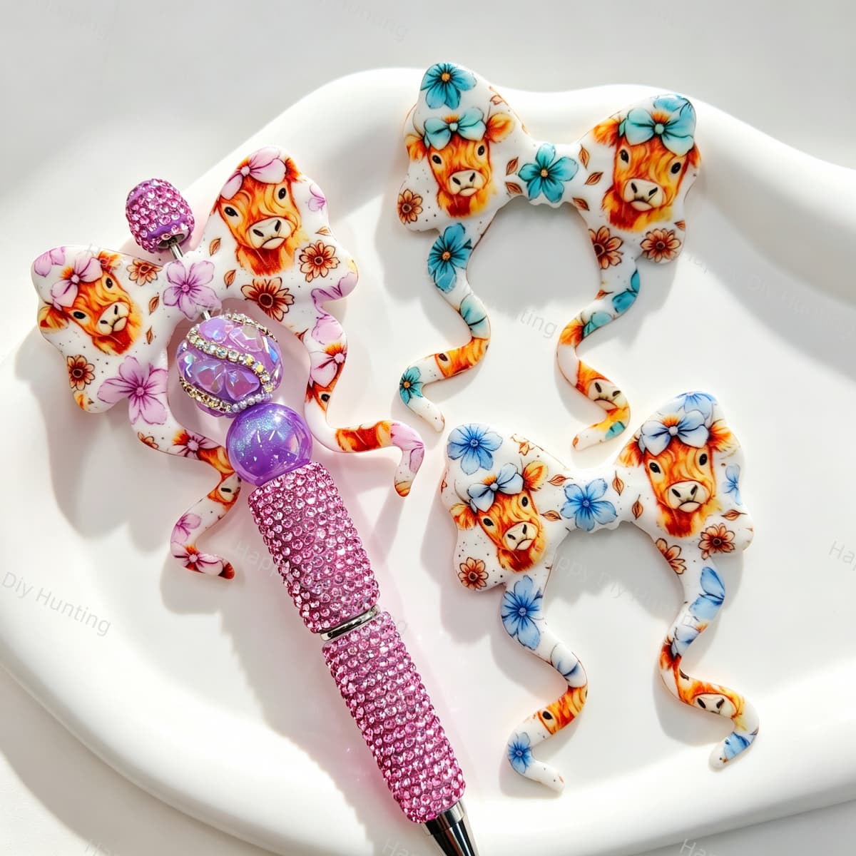 A collection of mixed color highland cow floral print silicone bow beads, including purple, blue, and teal designs, displayed with a pink rhinestone beadable pen. Soft, durable, and perfect for beadable pens, keychains, and handmade crafts.