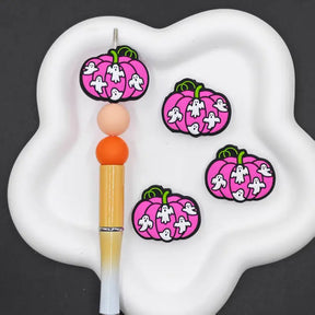 Three pink pumpkin-shaped charms with white ghosts and green stems are displayed on a white cloud-shaped tray next to a yellow-and-white pen on a dark gray background.