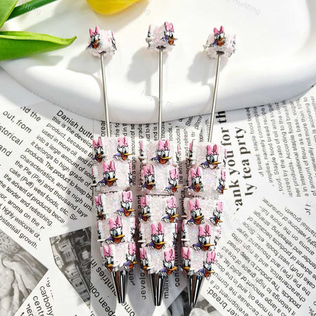 Three pink bow cartoon duck character beadable pens displayed on a newspaper background. Each pen features pink glitter beads decorated with white cartoon duck faces wearing pink bows, plus a silver metal tip. Perfect for DIY crafts, focal beads, and handmade gifts.
