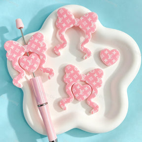 Bow-shaped silicone focal beads featuring a pink monogram V*L pattern design. Soft, durable, and flexible, perfect for beadable pens, keychains, and handmade crafts.