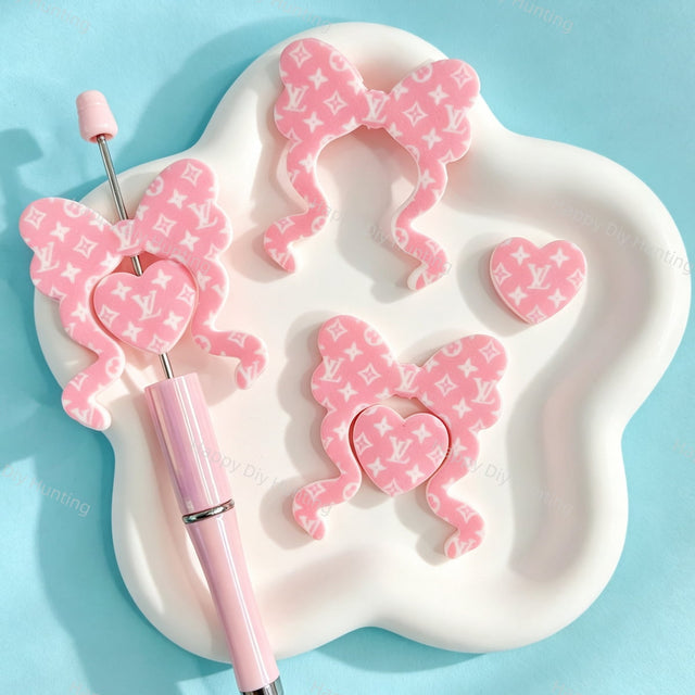 Bow-shaped silicone focal beads featuring a pink monogram V*L pattern design. Soft, durable, and flexible, perfect for beadable pens, keychains, and handmade crafts.