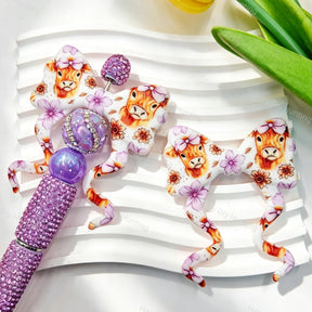 Purple highland cow floral print silicone bow beads displayed with a purple rhinestone beadable pen. Featuring an adorable highland cow with a bow and purple flowers. Soft, durable, and perfect for beadable pens, keychains, and handmade crafts.