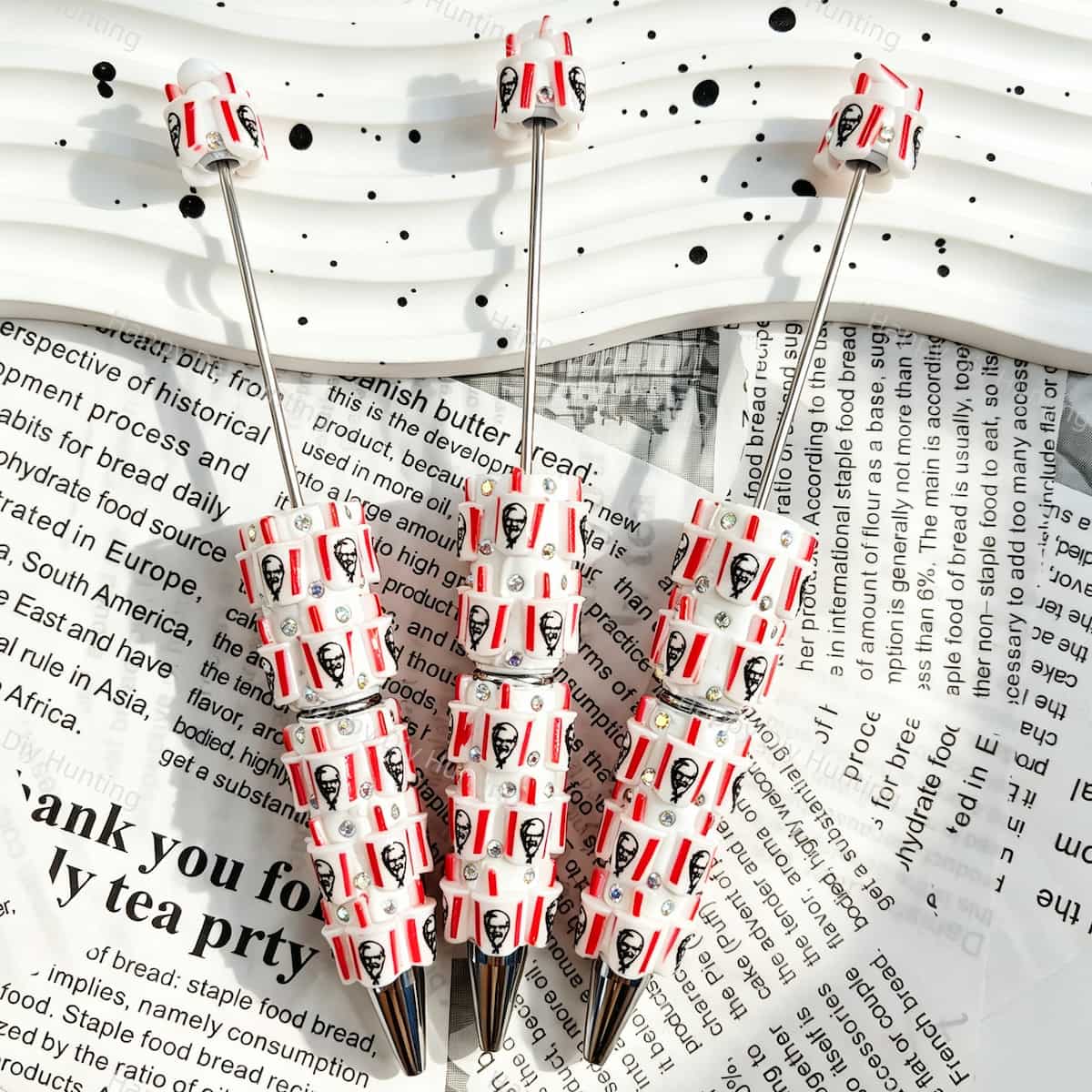 Three red and white elderly figure print beadable pens displayed on a newspaper background. Each pen features white beads decorated with red and white stripes, elderly figure faces, and colorful rhinestones, plus a silver metal tip. Ideal for DIY crafts, focal beads, and handmade gifts.
