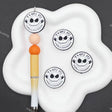 A white cloud-shaped pen stand holding a decorative pen with orange and peach beads, alongside four round stickers featuring a black-and-white skull face and the text 'THIS IS HALLOWEEN' on a dark gray background.