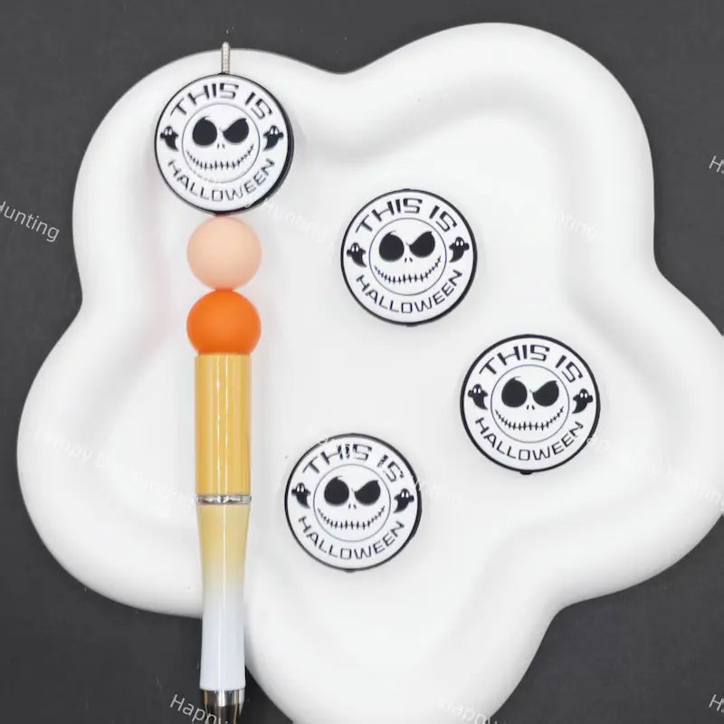 A white cloud-shaped pen stand holding a decorative pen with orange and peach beads, alongside four round stickers featuring a black-and-white skull face and the text 'THIS IS HALLOWEEN' on a dark gray background.