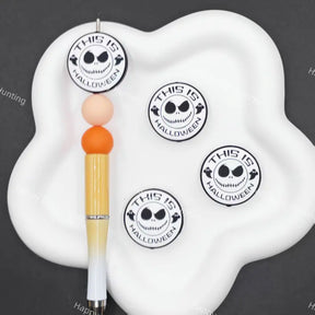 A white cloud-shaped pen stand holding a decorative pen with orange and peach beads, alongside four round stickers featuring a black-and-white skull face and the text 'THIS IS HALLOWEEN' on a dark gray background.