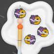 Four Halloween-themed rubber band charms featuring yellow pumpkins with purple flowers and a banner reading 'SCOURGE THE STICKIN' GUM' are displayed on a white cloud-shaped stand next to a wooden pen.