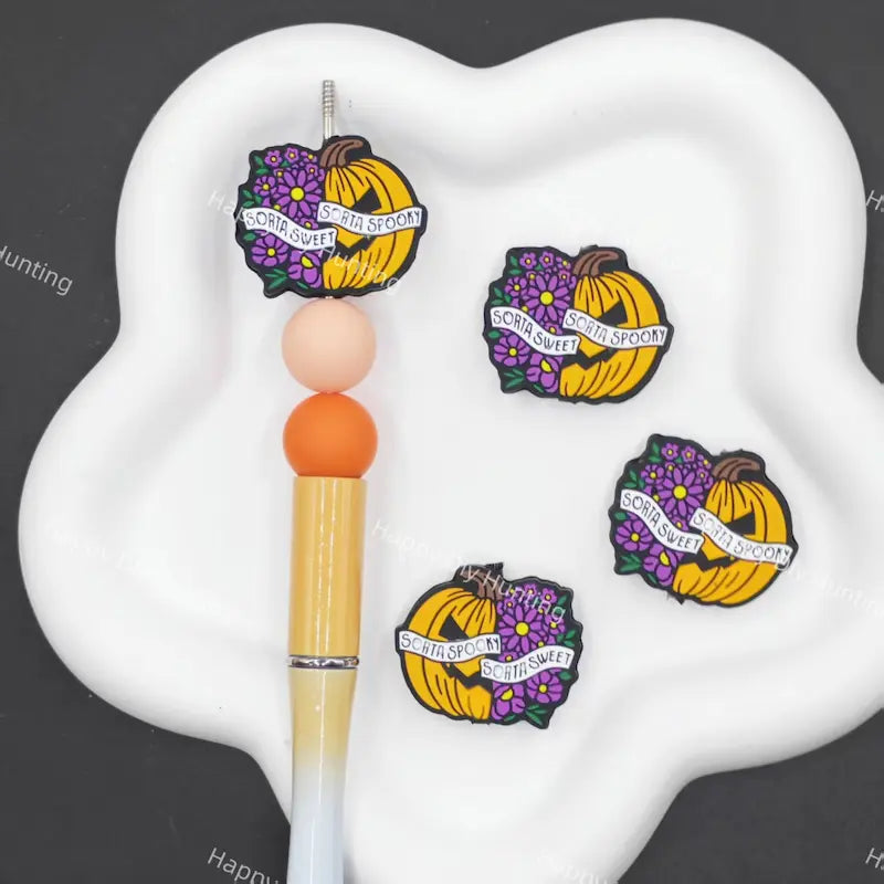 Four Halloween-themed rubber band charms featuring yellow pumpkins with purple flowers and a banner reading 'SCOURGE THE STICKIN' GUM' are displayed on a white cloud-shaped stand next to a wooden pen.