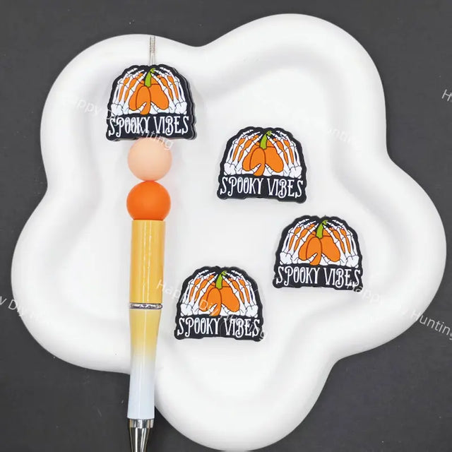 Three identical 'Spooky Vibes' pumpkin patch enamel pins are displayed on a white cloud-shaped tray next to a yellow and white pen, set against a dark gray background.
