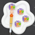 Three small decorative stickers reading 'THE BOO CREW' in purple, orange, and green are shown on a white cloud-shaped holder next to a yellow-and-white pen. The background is dark gray.