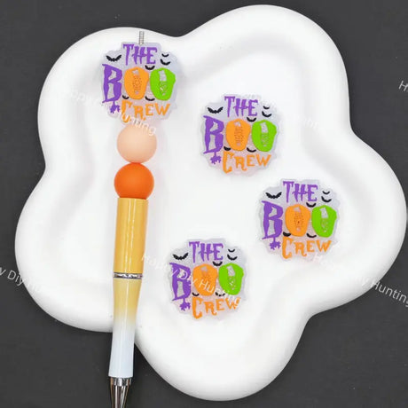 Three small decorative stickers reading 'THE BOO CREW' in purple, orange, and green are shown on a white cloud-shaped holder next to a yellow-and-white pen. The background is dark gray.