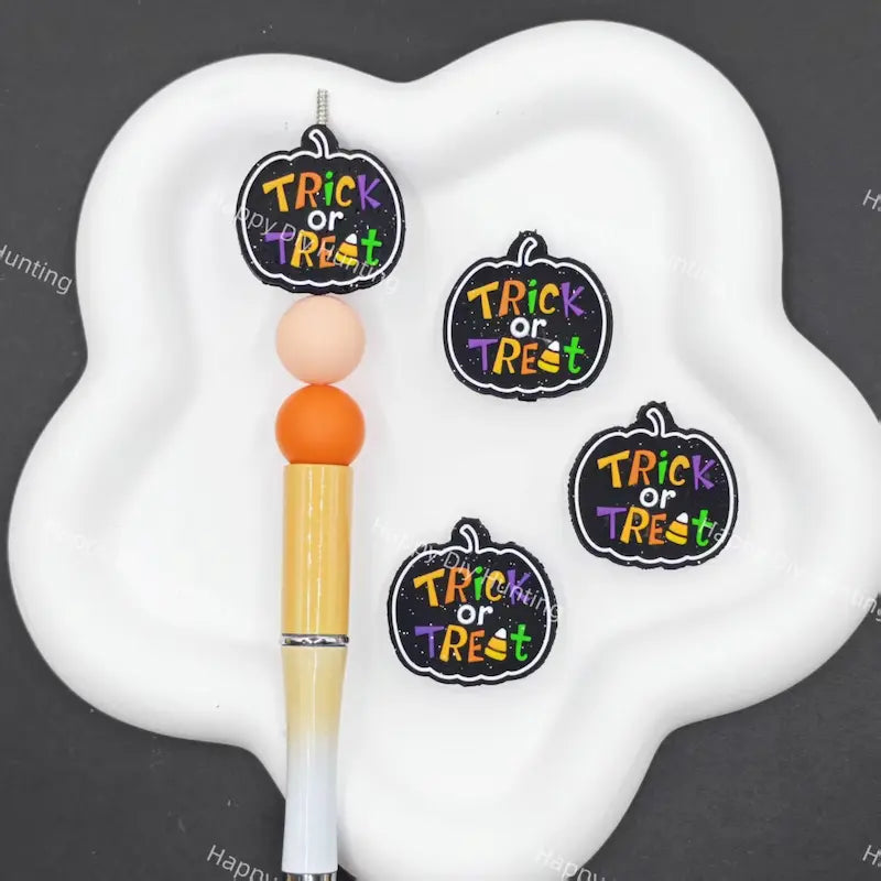 Three pumpkin-shaped 'Trick or Treat' stickers displayed on a white cloud-shaped tray next to a yellow and white pen, set against a dark gray background.