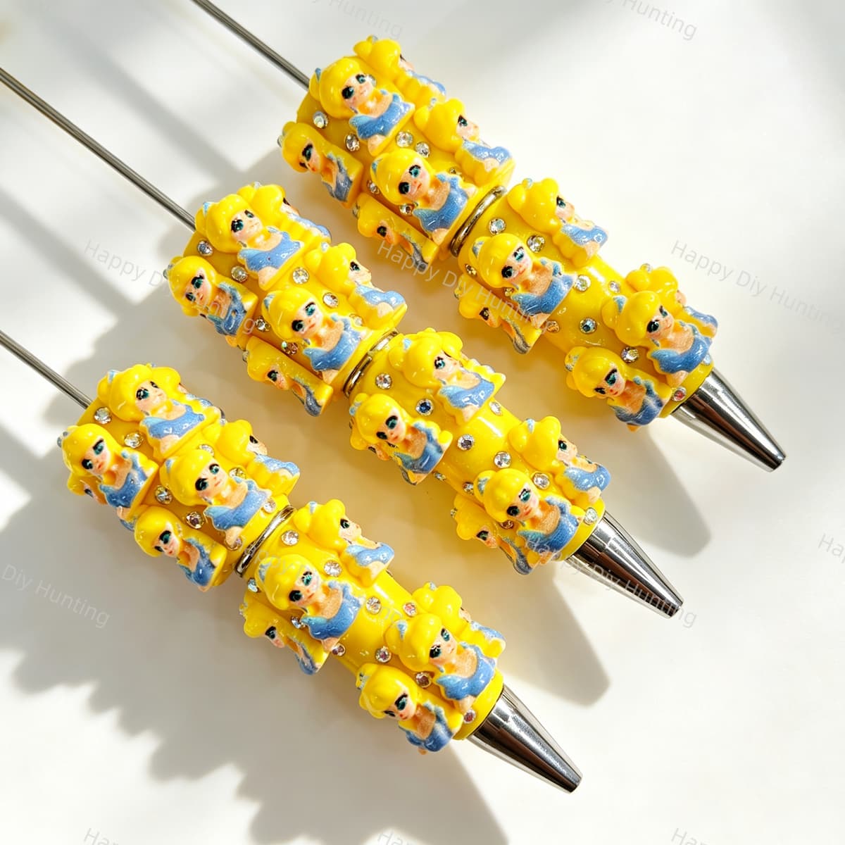Close-up of four yellow haired cartoon girl character beadable pens on a white background. Each pen features yellow beads decorated with cartoon girl faces wearing blue dresses and clear rhinestones, paired with silver metal tips. Ideal for DIY beadable pen projects, focal beads, and handmade crafts.