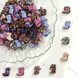 A pile of small cowboy boot charms or beads in assorted colors and patterns, arranged on a white surface with a scalloped glass dish and several individual boots displayed around it.