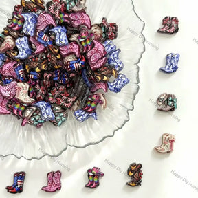 A pile of small cowboy boot charms or beads in assorted colors and patterns, arranged on a white surface with a scalloped glass dish and several individual boots displayed around it.